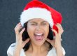 Closeup portrait of a cute Christmas woman with a red Santa Claus hat white dress screaming out loud frustrated eyes shut in rage. Negative human emotion on an isolated grey background.