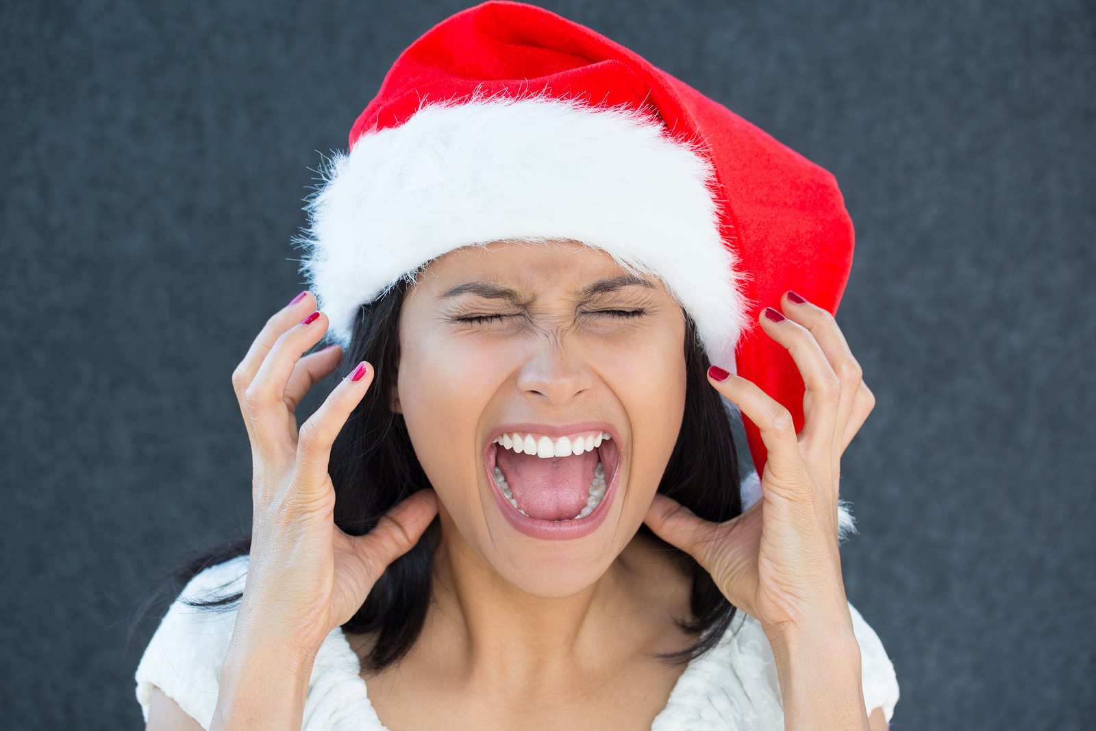 Closeup portrait of a cute Christmas woman with a red Santa Claus hat white dress screaming out loud frustrated eyes shut in rage. Negative human emotion on an isolated grey background.