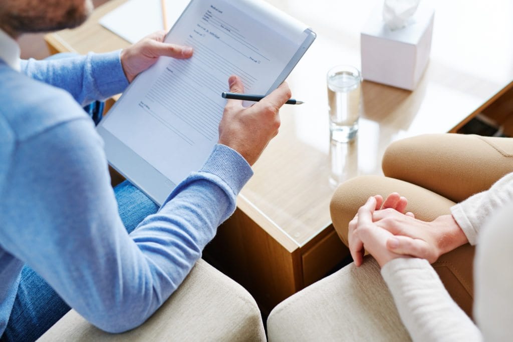 Mature psychiatrist sitting next to his female patient and asking her questions necessary for filling in medical card, close-up shot